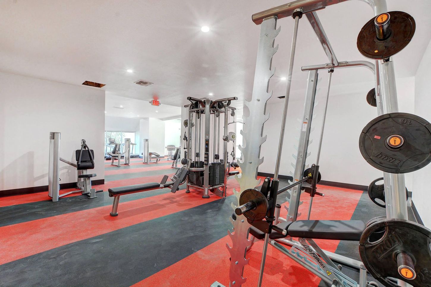 Gym interior with weight machines, a bench, and weights. Red and black flooring. Brightly lit space.