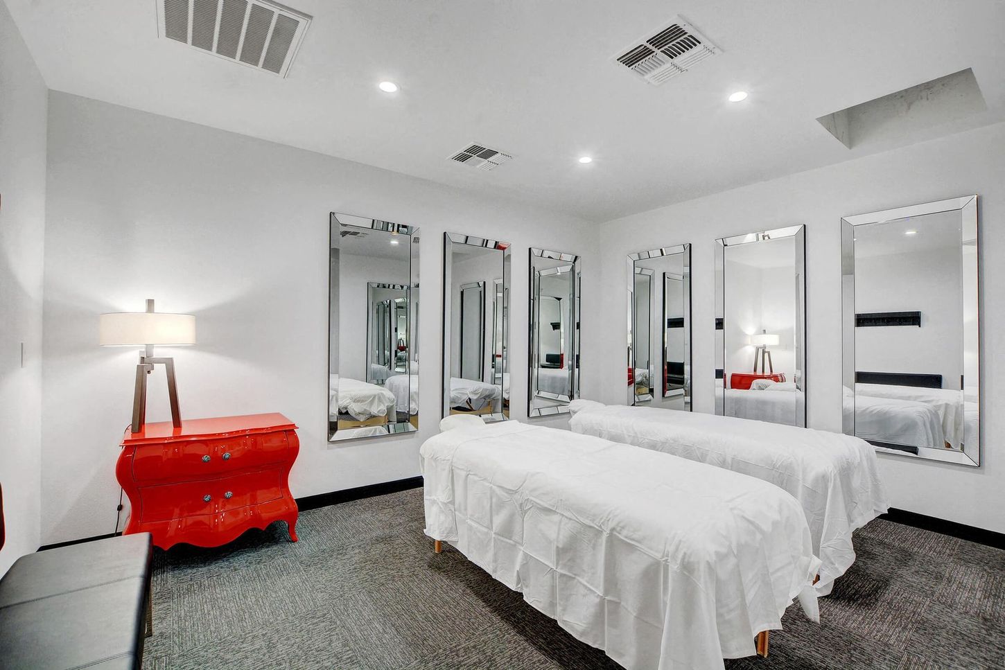 Two massage tables in a white room with mirrors, red cabinet, and overhead lights.