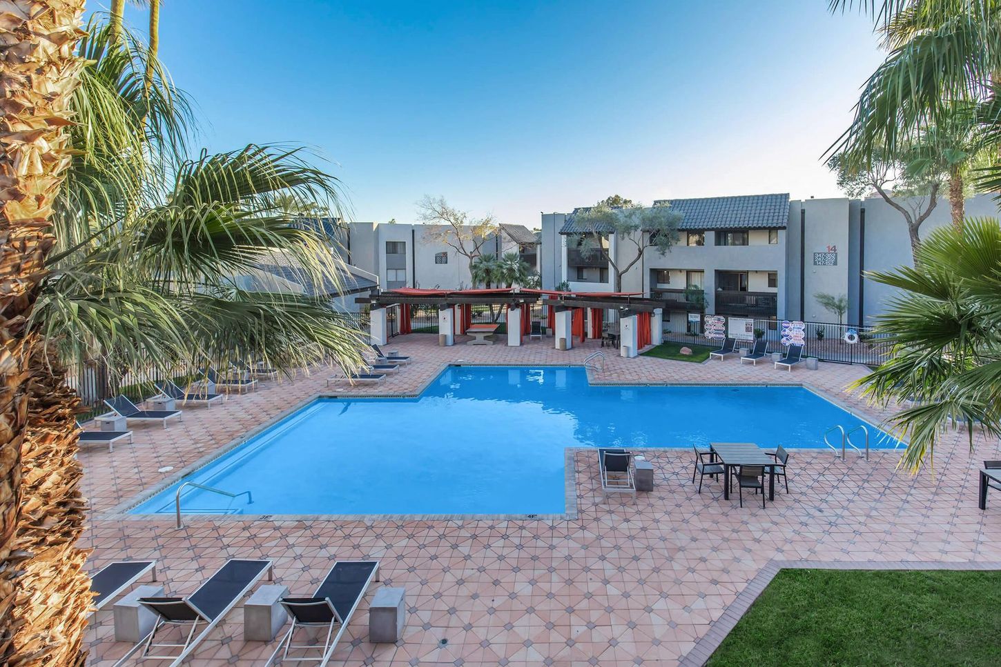 Pool surrounded by lounge chairs, tables, and palm trees. Apartment buildings in background under a blue sky.