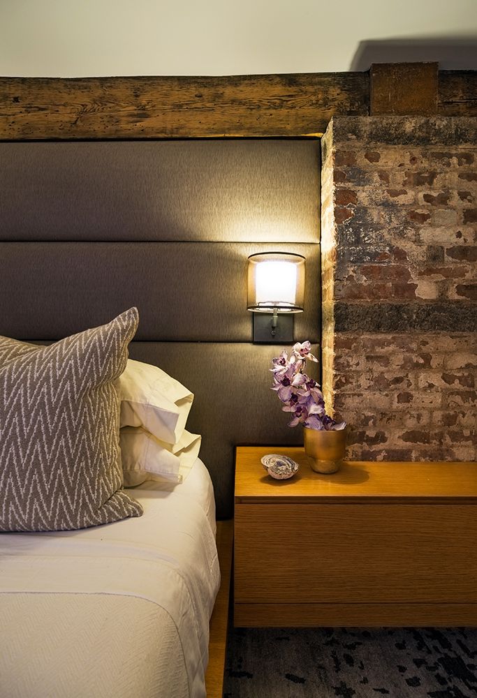 A bedroom with a bed , nightstand , lamp and brick wall.