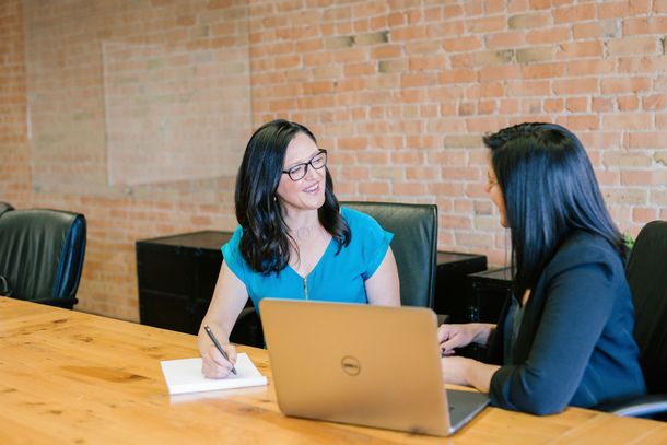Two women at a wooden table; one writes in a notepad, the other uses a laptop in a room with brick walls.