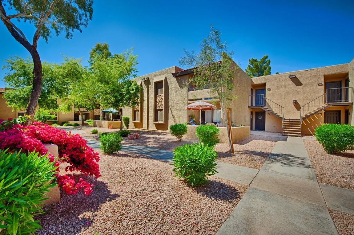 Desert-style apartment courtyard with stone buildings, gravel paths, and bright pink flowering shrubs.