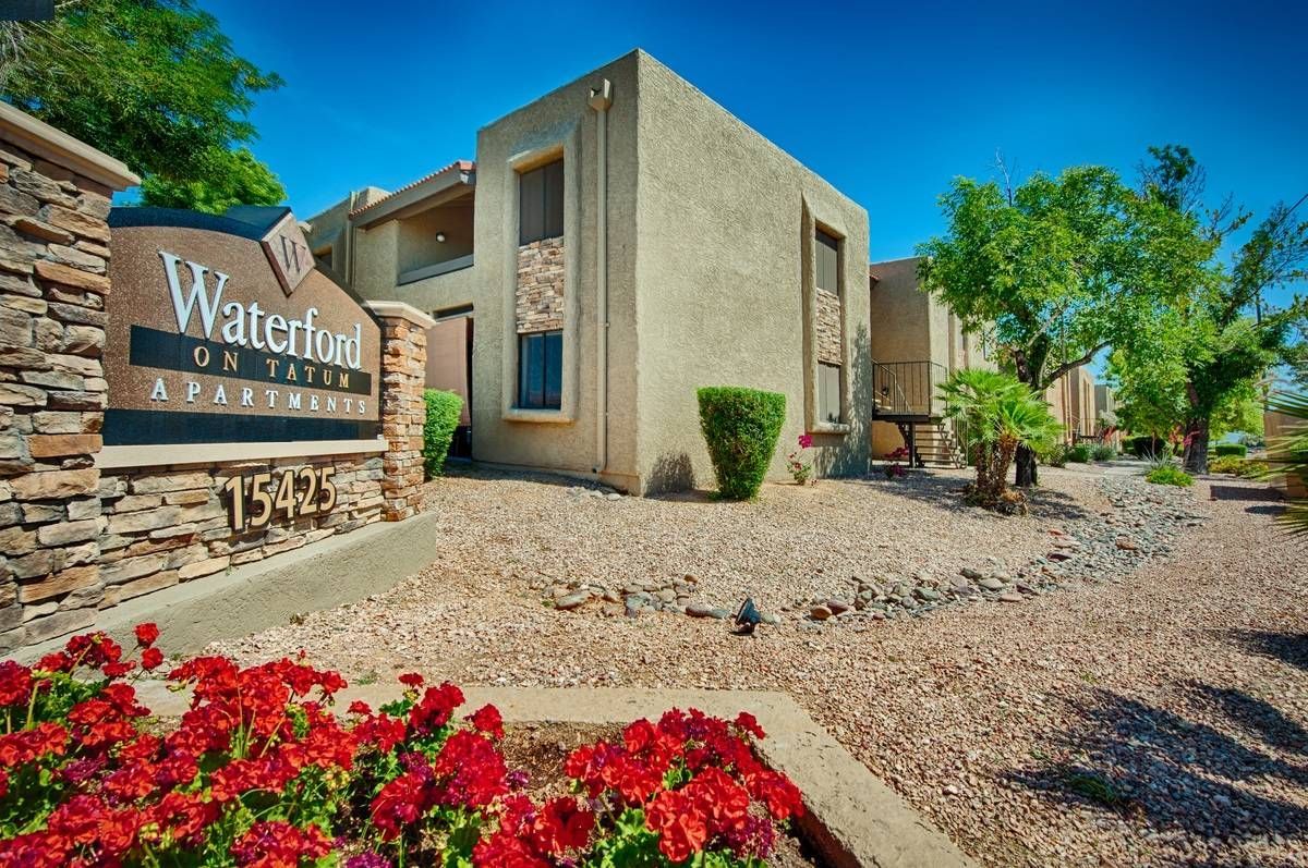 Apartment complex entrance with stone sign, desert landscaping, and red flowers under a blue sky