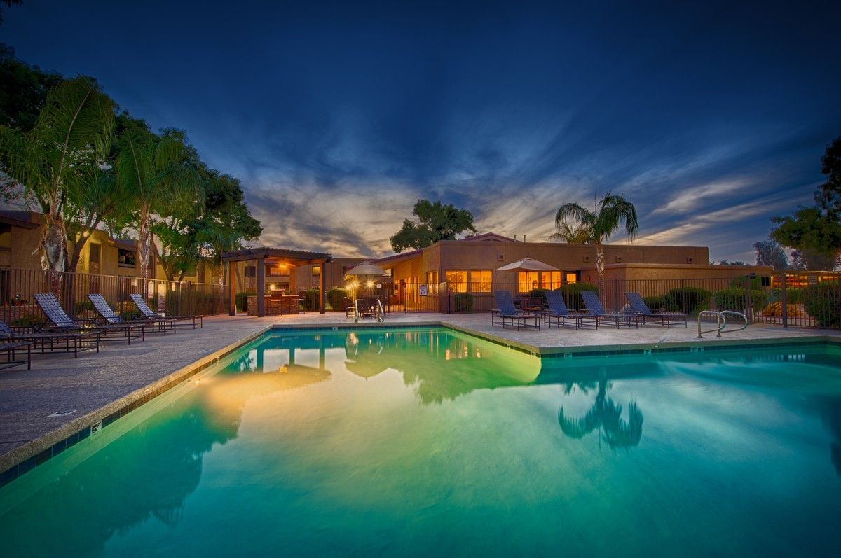 Lit resort pool at dusk with turquoise water, lounge chairs, and warm glowing buildings in the background