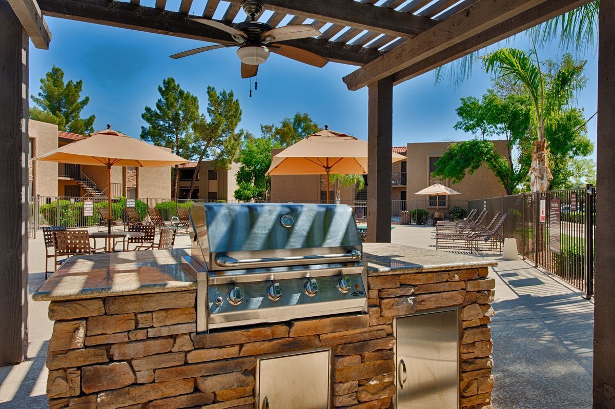 Outdoor patio with a stone grill, pergola shade, tables, and orange umbrellas by a pool