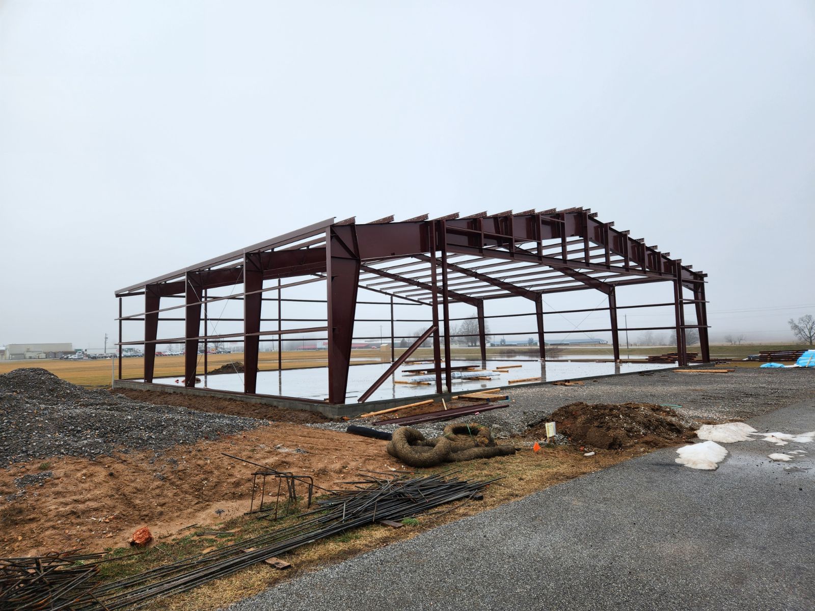 Steel frame of a building under construction on a cloudy day; wet ground and gravel in the foreground.