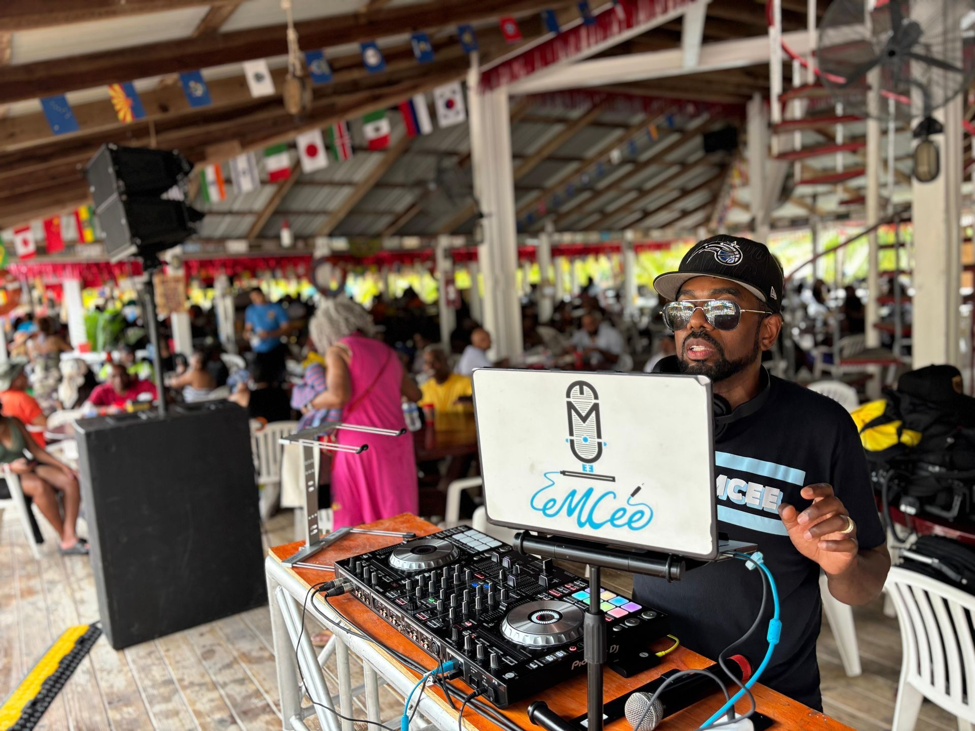 A DJ wearing sunglasses and a black baseball cap operates a mixing console at a table in a busy, open-air pavilion.