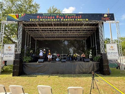 A large stage at a Heritage Day Festival for First Missionary Baptist Church, with people sitting under a truss canopy.