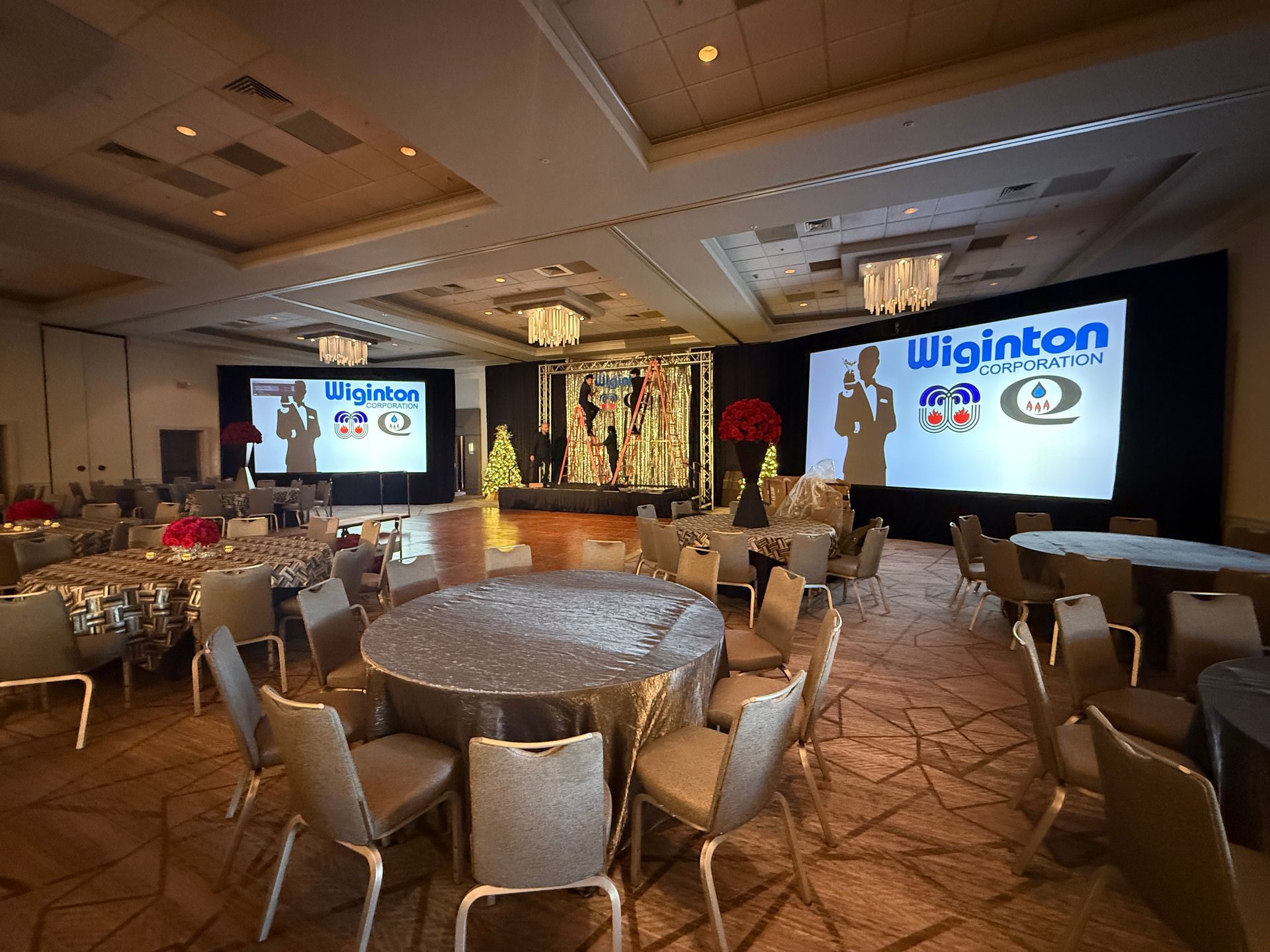 A large hotel ballroom set for a banquet with round tables, chairs, and two screens displaying Wiginton branding.