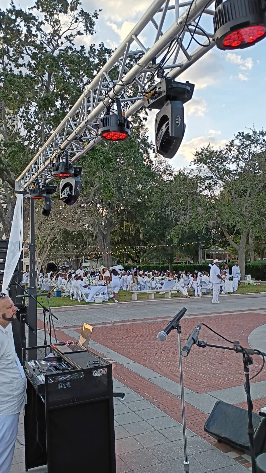 Outdoor event with a stage lighting truss, audio equipment, and people in white clothing seated in a park setting.