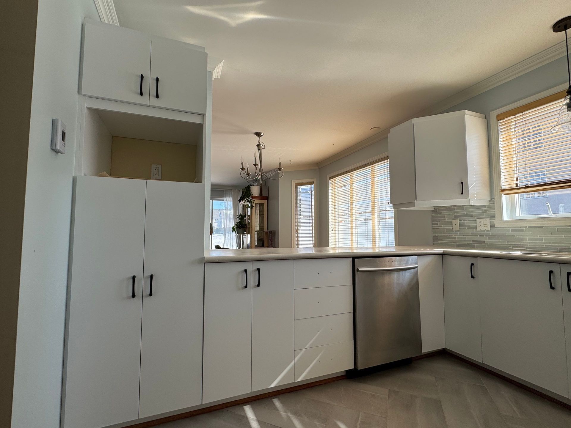 Cuisine équipée d'armoires blanches avec poignées noires, lave-vaisselle, plan de travail et fenêtre avec stores.