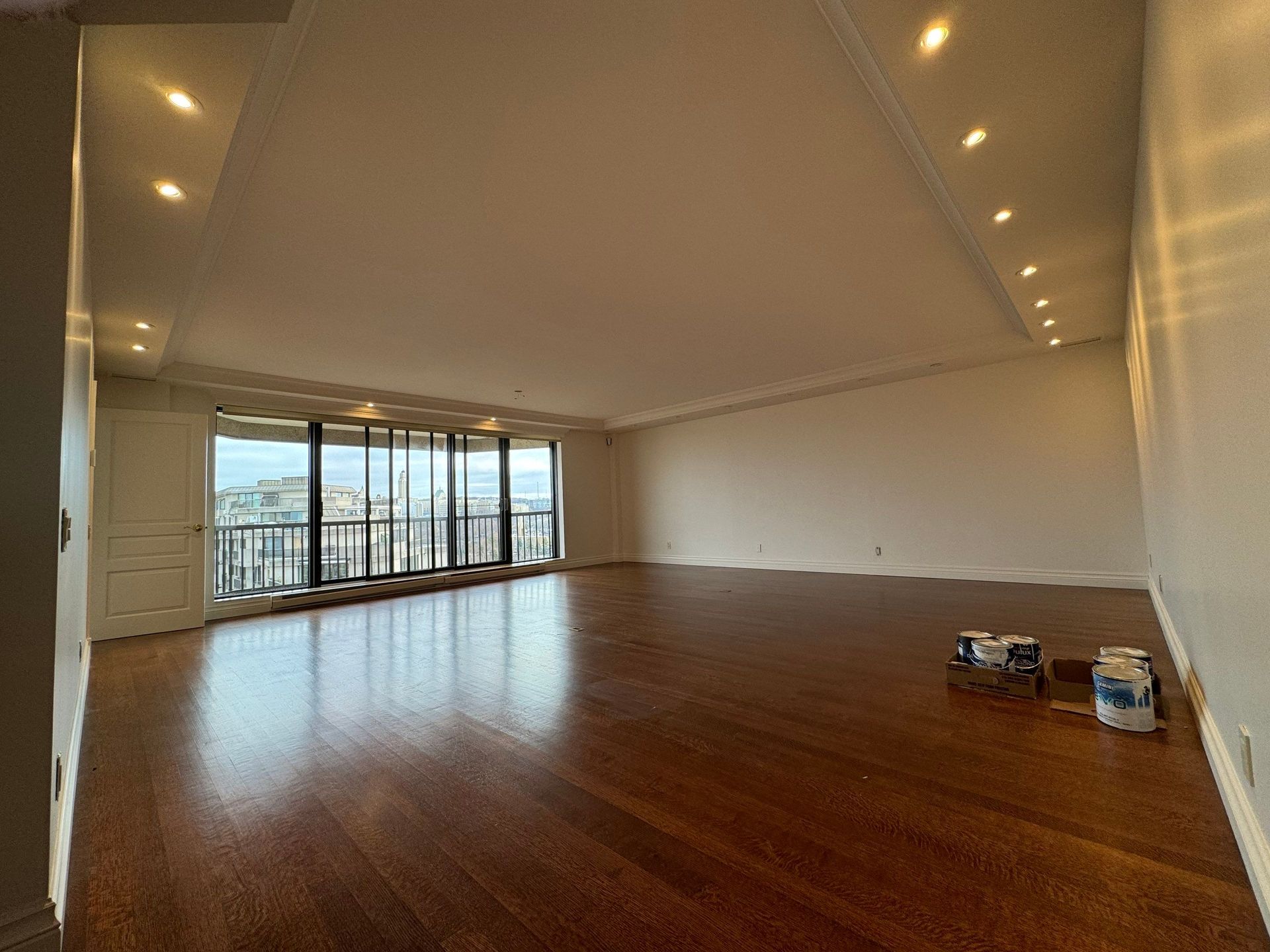 Salon vide avec parquet, grandes fenêtres, éclairage encastré et vue sur la ville.