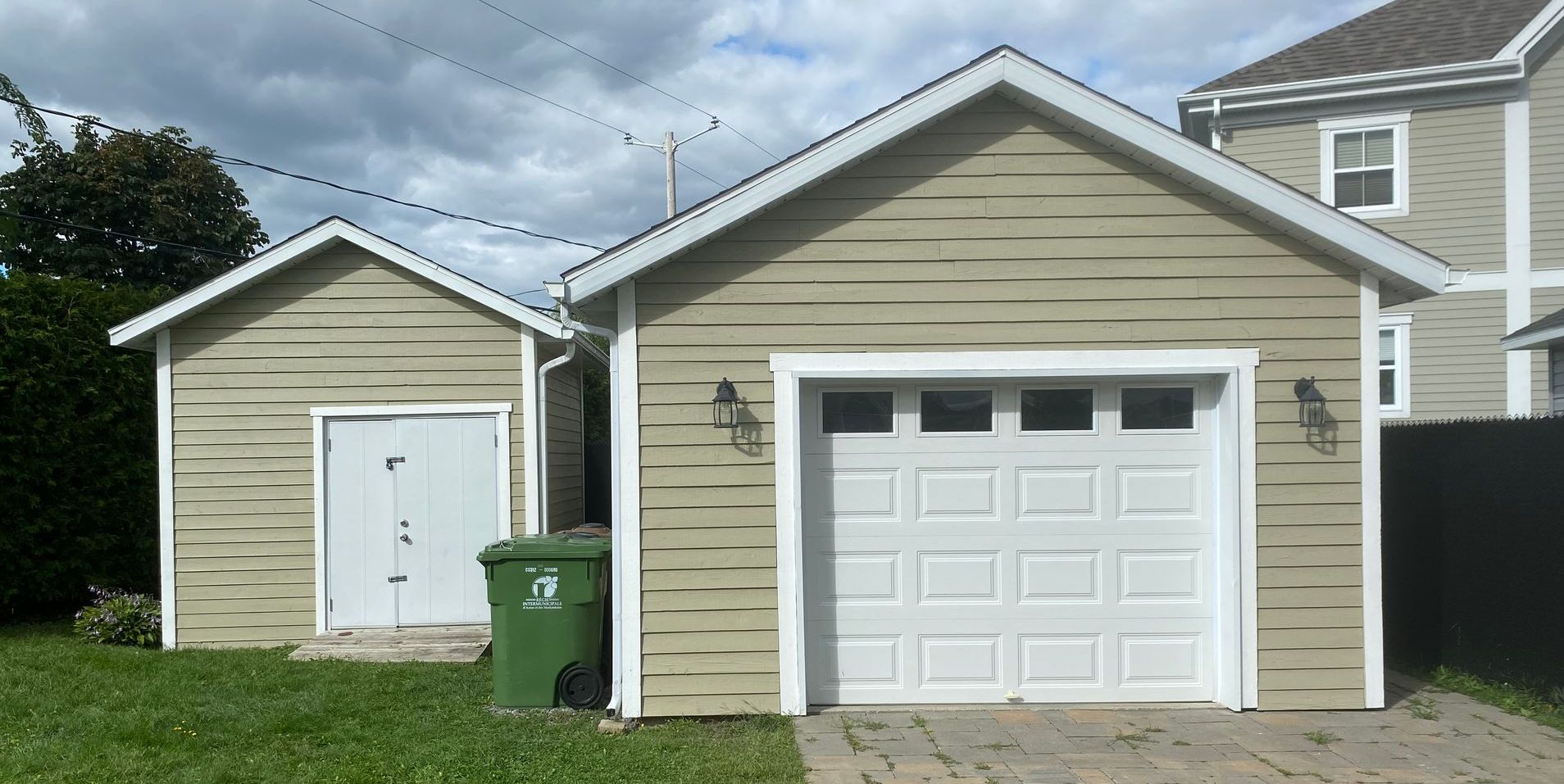 Deux bâtiments beige clair avec des portes blanches et une porte de garage. Une poubelle verte se trouve devant.