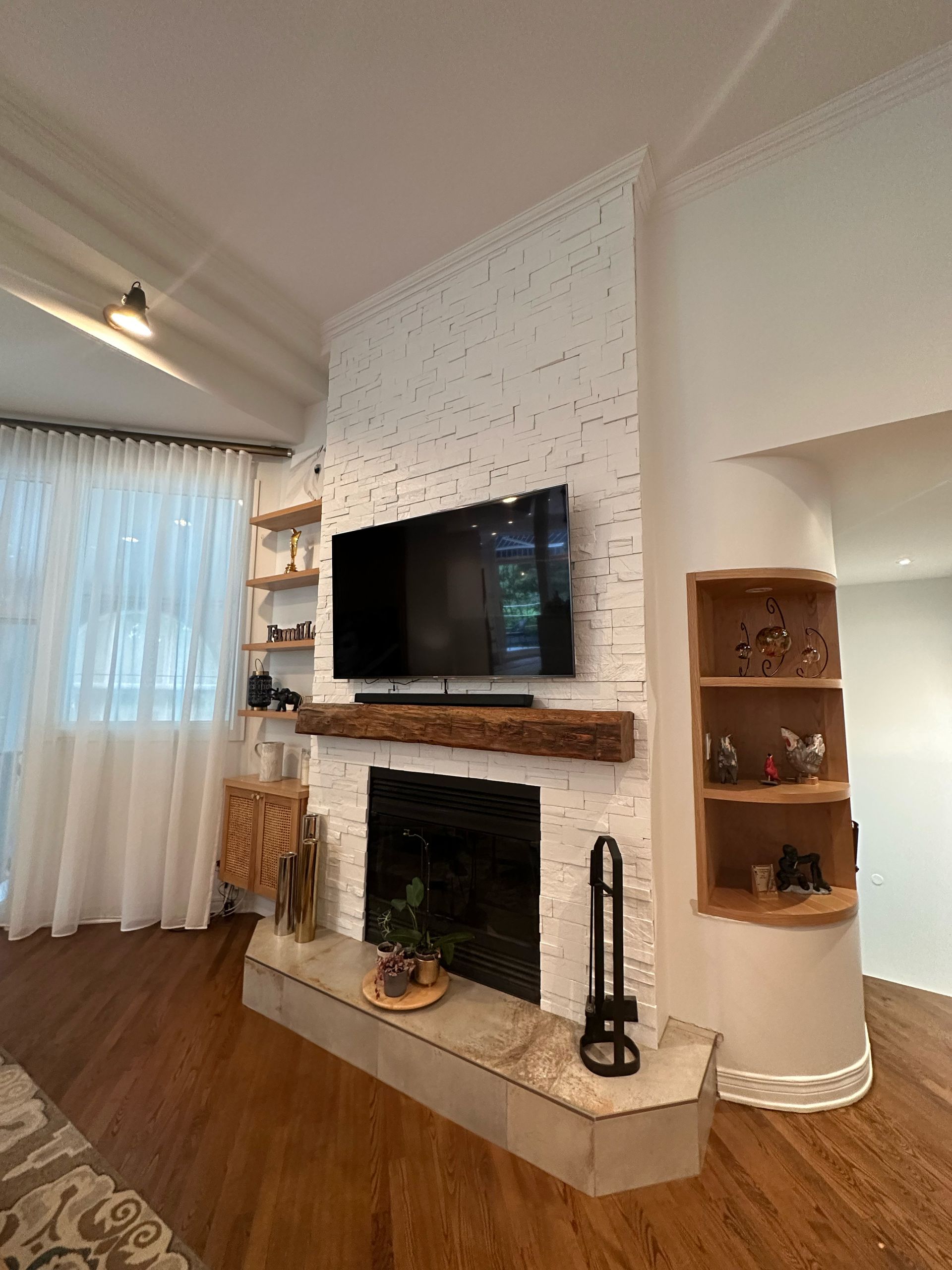 Salon avec cheminée en briques blanches, télévision et étagères en bois. Parquet et voilages.