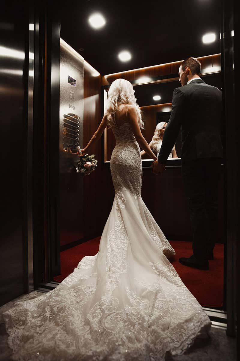 Bride and groom in an elevator. She wears a white gown with a long train, holding a bouquet, and he wears a suit.