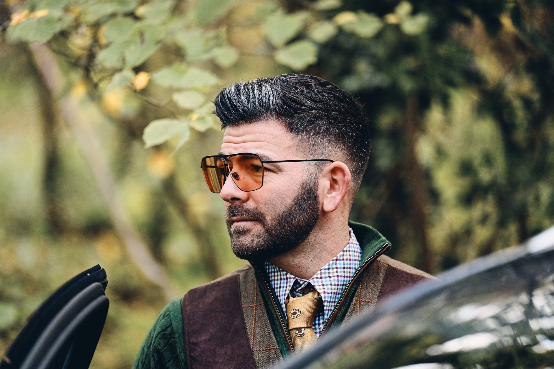 Man with beard and orange tinted glasses looking to the side outdoors, wearing a vest and collared shirt.