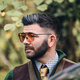 Man with beard, wearing sunglasses, shirt, and vest outdoors.