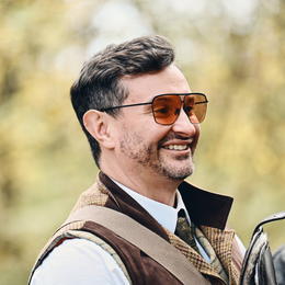 Man with sunglasses, smiling, wearing a vest and tie, outdoors.