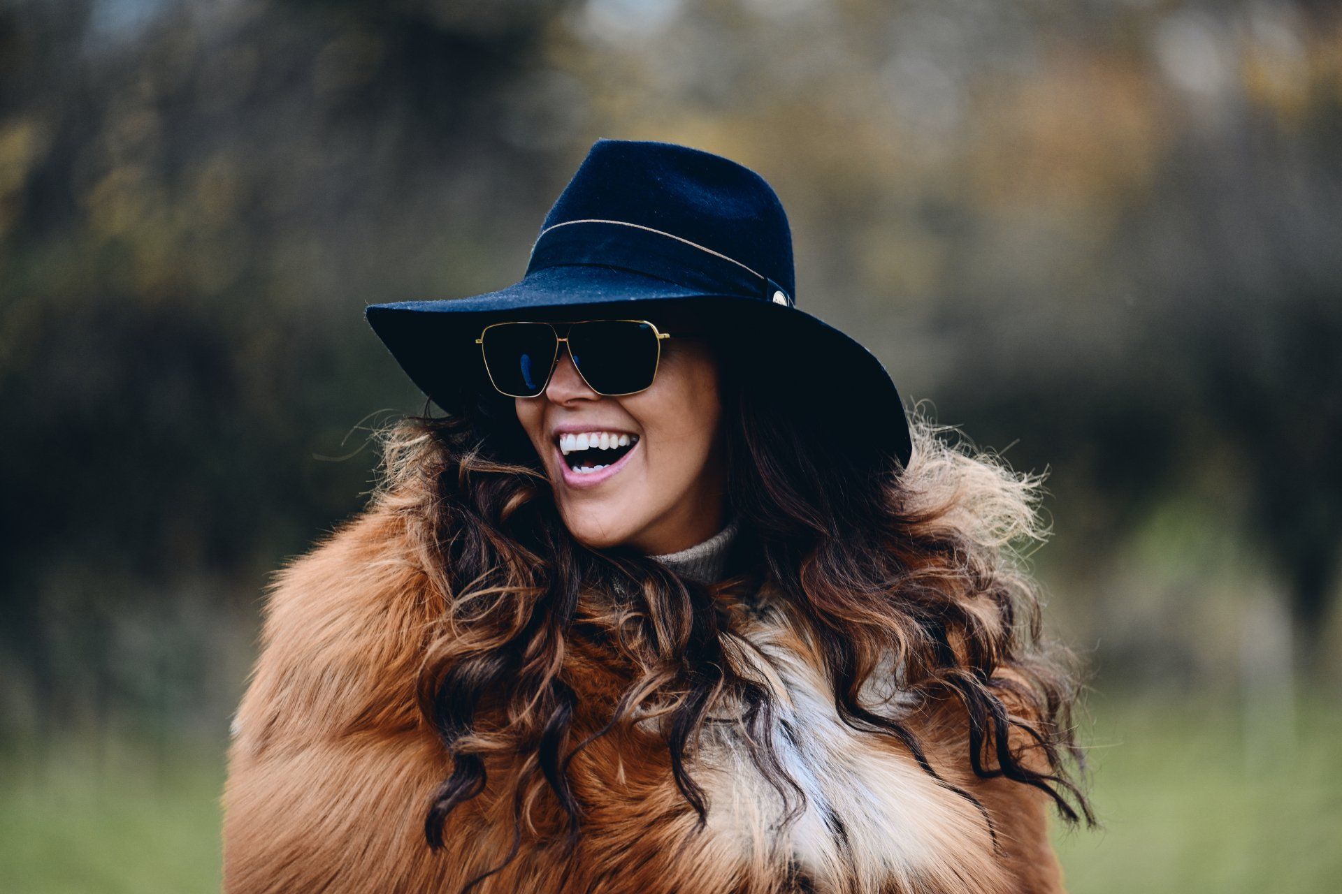 Woman in a black hat and sunglasses, laughing outdoors, wearing a furry brown coat.