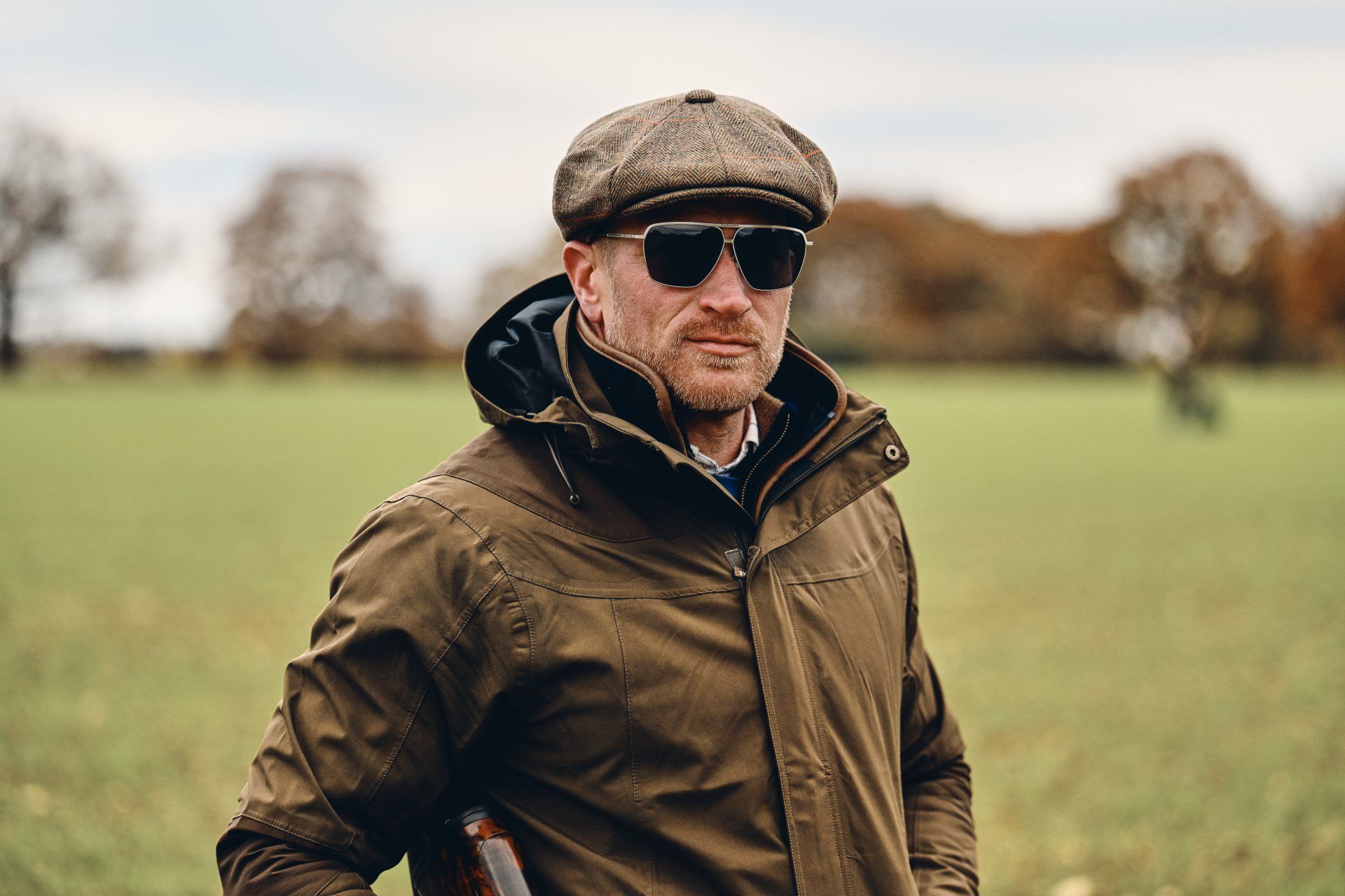 Man in cap and sunglasses, wearing jacket, stands in field with trees.