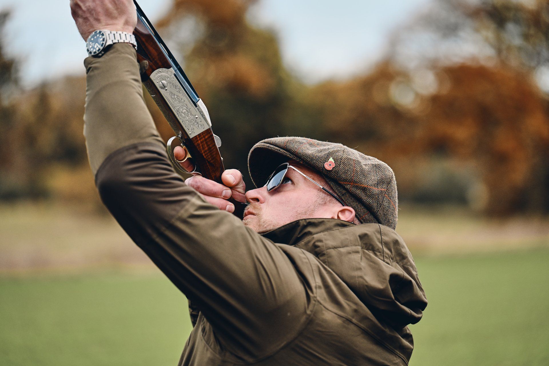 Man aiming a shotgun upwards, wearing a cap and sunglasses, outdoors on a grassy field.