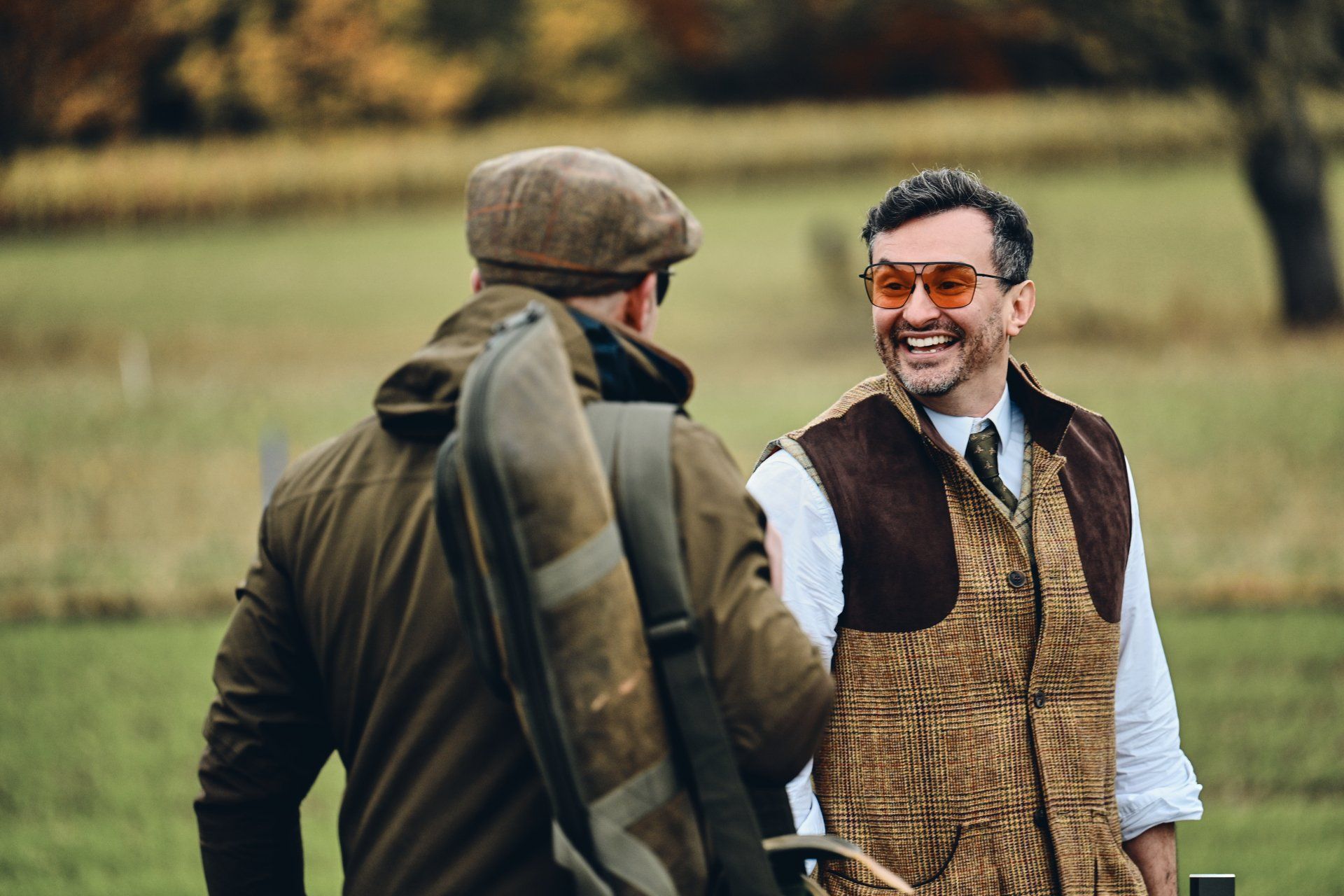 Two men talking outdoors, one with shotgun case, other wearing vest and orange sunglasses, smiling.