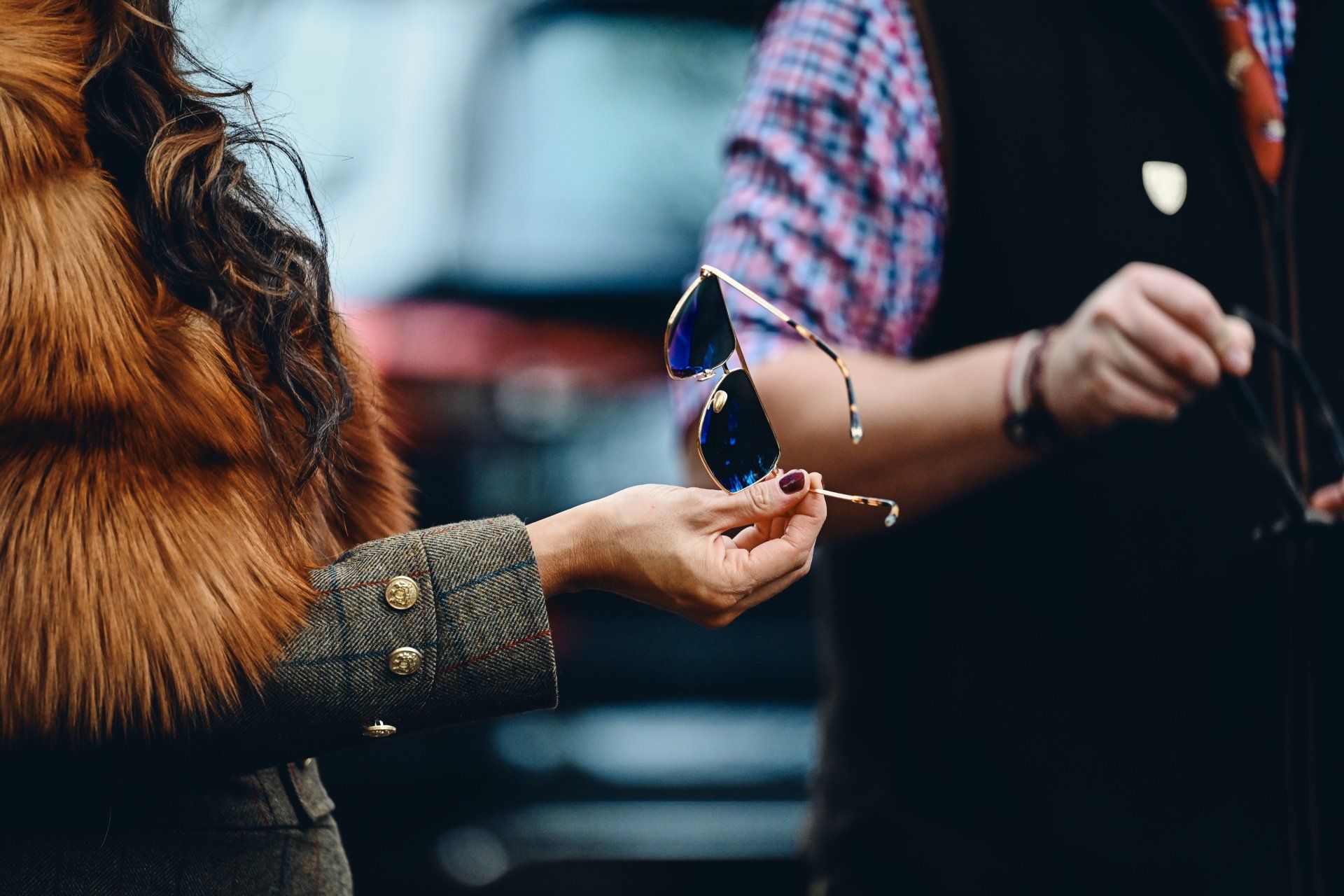 Woman in faux fur vest holding sunglasses, person in vest nearby.