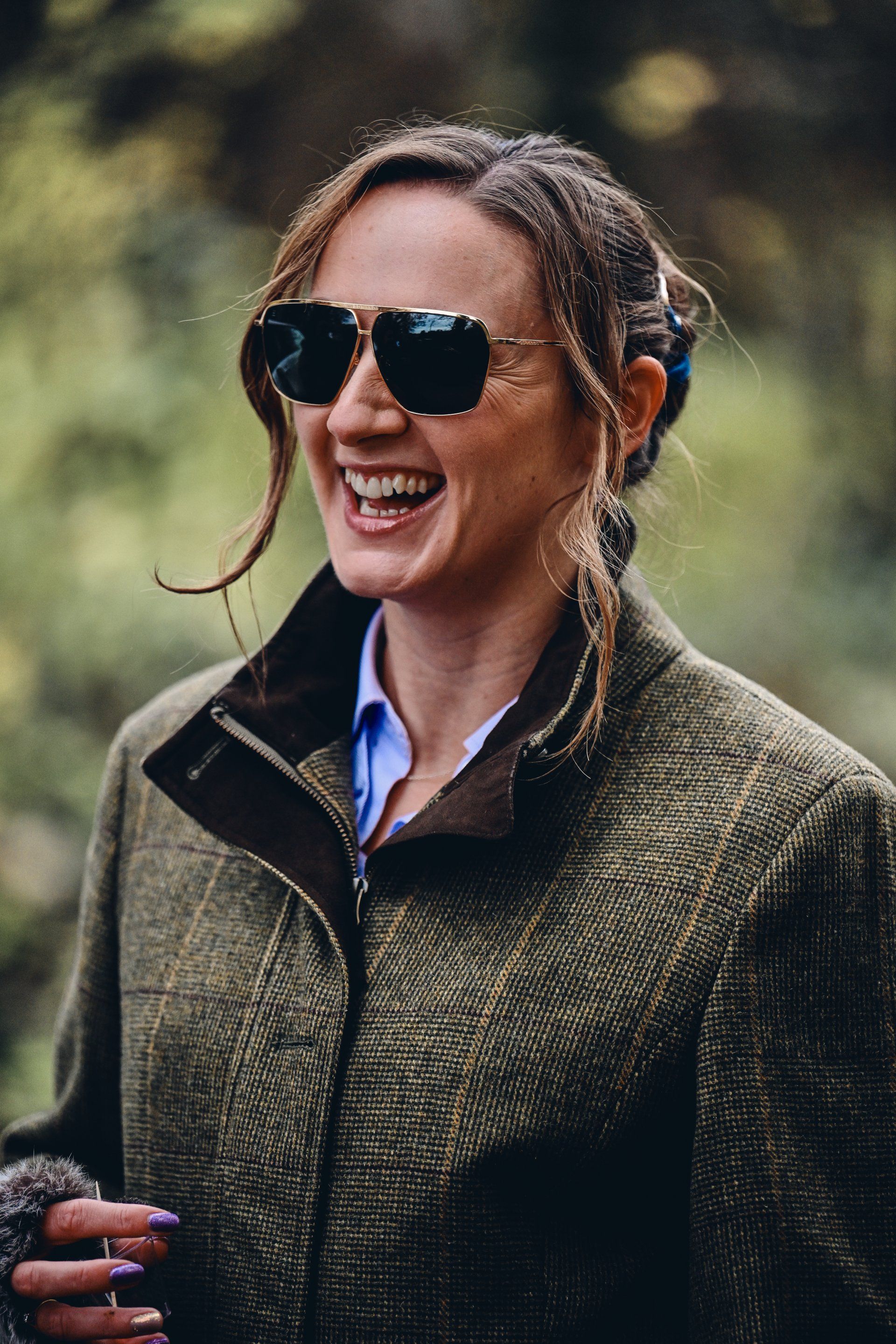 Woman laughing, wearing sunglasses and a tweed jacket, outdoors.