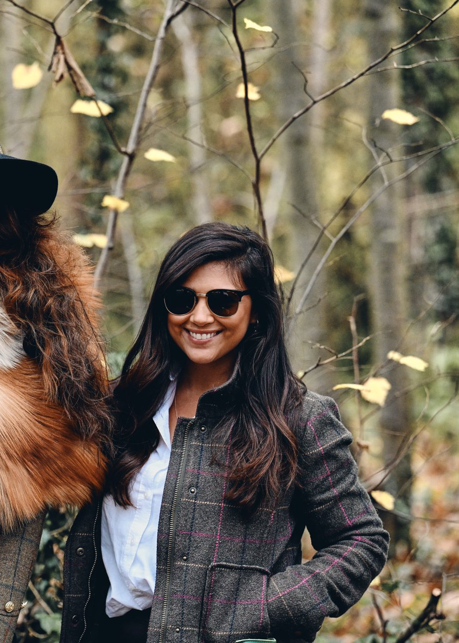 Woman in sunglasses and tweed jacket smiles in a wooded area.