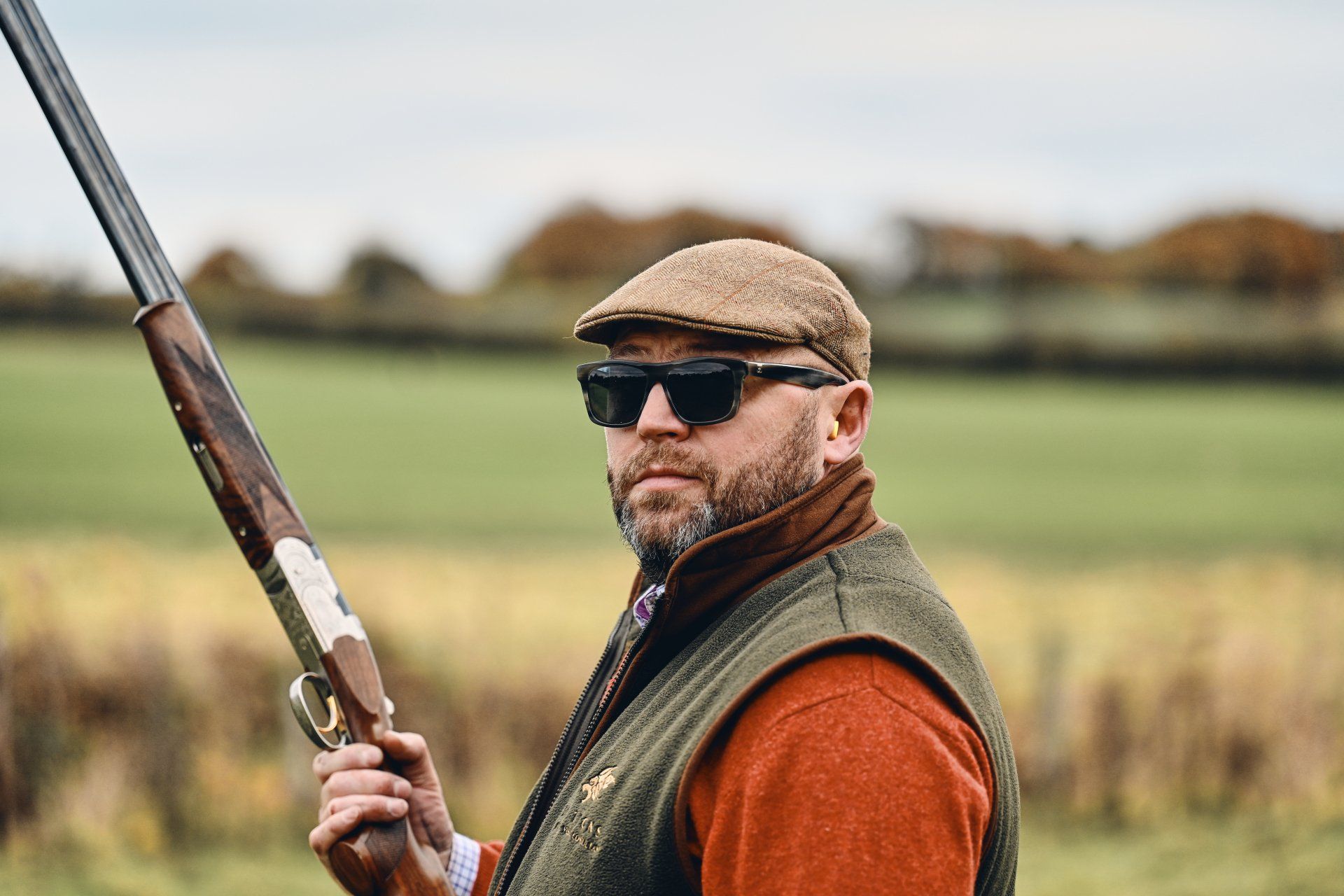 Man in tweed cap, sunglasses, and vest holding a shotgun outdoors in a field.