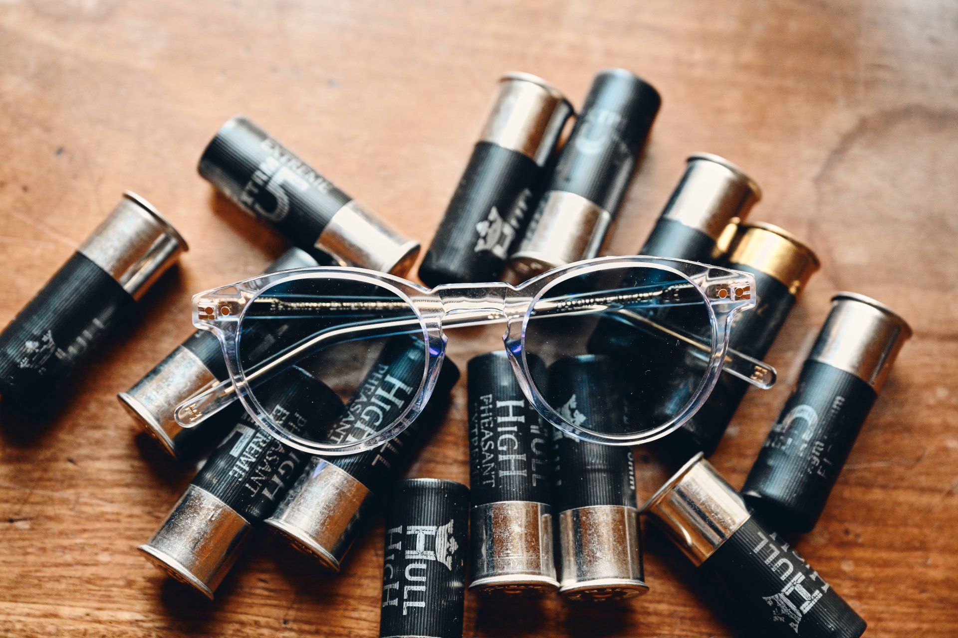 Clear-framed sunglasses resting on a pile of black and silver shotgun shells on a wooden surface.