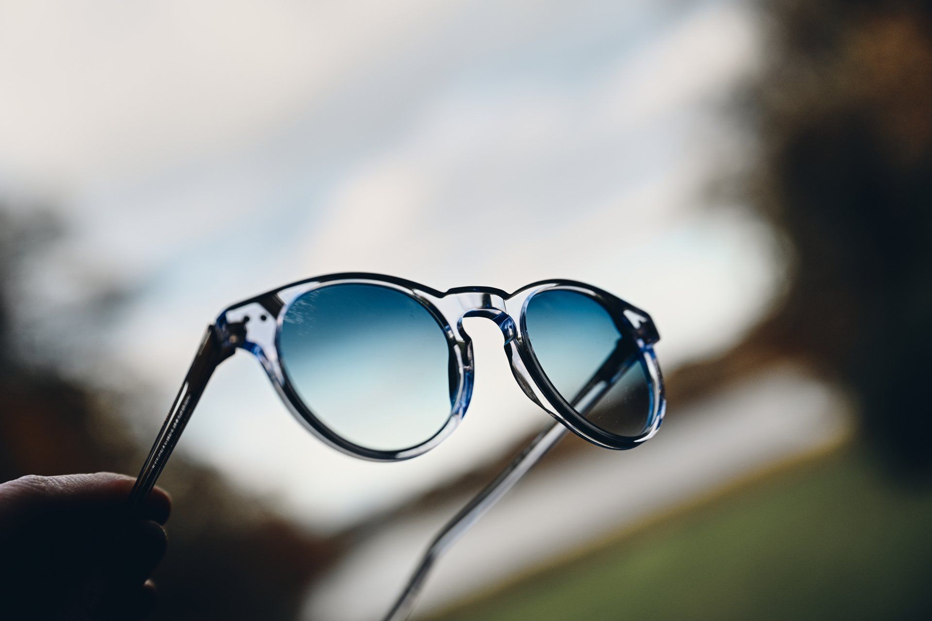 Sunglasses held up against a blurry background, clear frames with blue-tinted lenses.