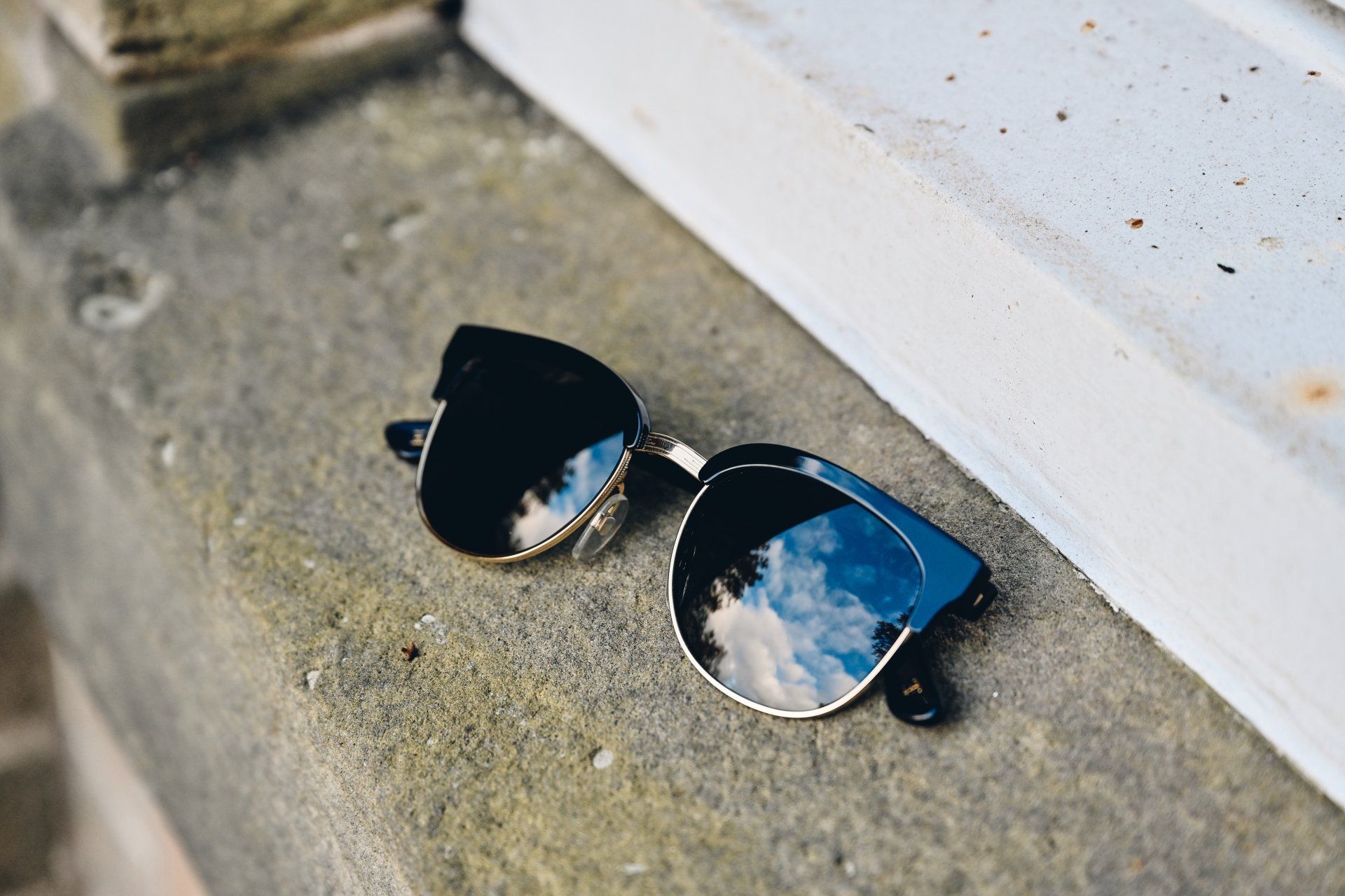 Sunglasses with mirrored lenses on a stone ledge, reflecting a cloudy sky.