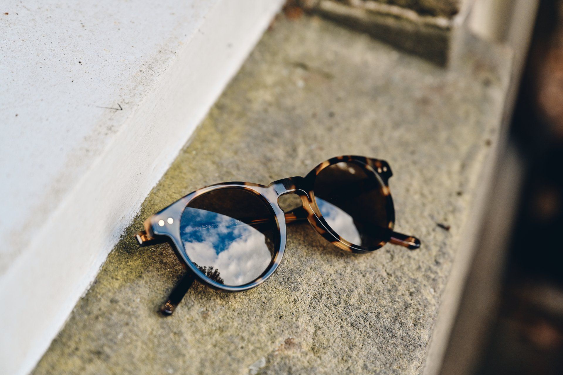 Sunglasses with tortoise-shell frames reflect a cloudy blue sky, resting on a stone ledge.