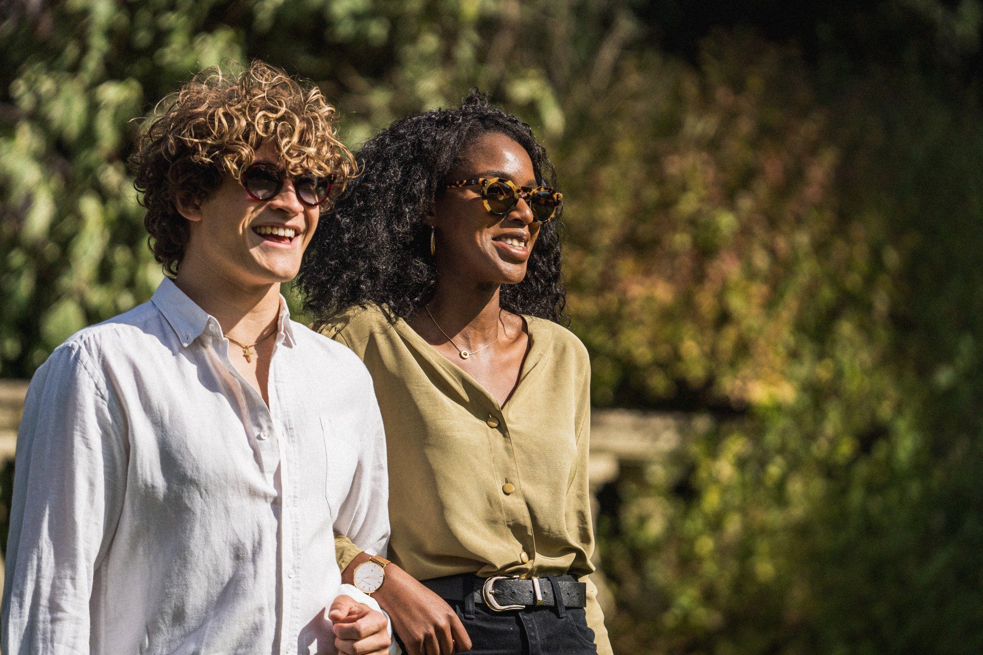 Two people smiling, wearing sunglasses, walking outdoors. Sunny day, greenery in the background.
