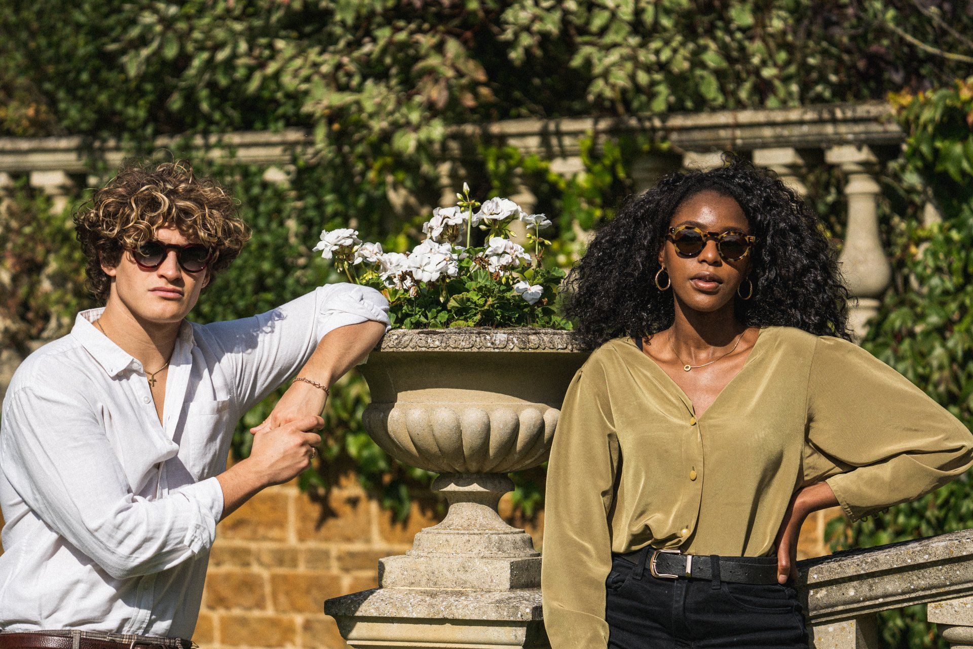 Two people wearing sunglasses pose near a stone planter outdoors.