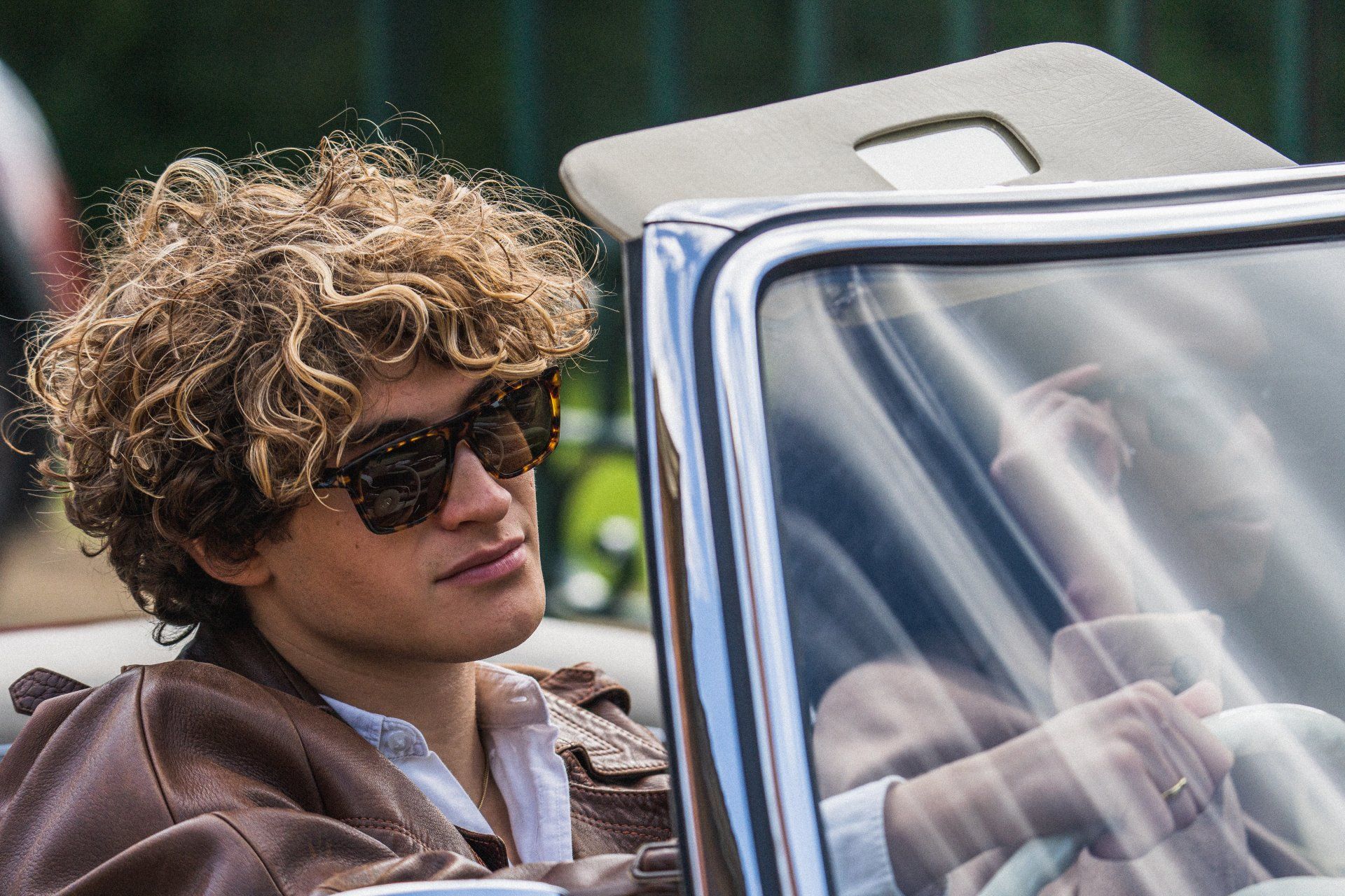 Man with curly hair and sunglasses in a classic car, looking to the side.