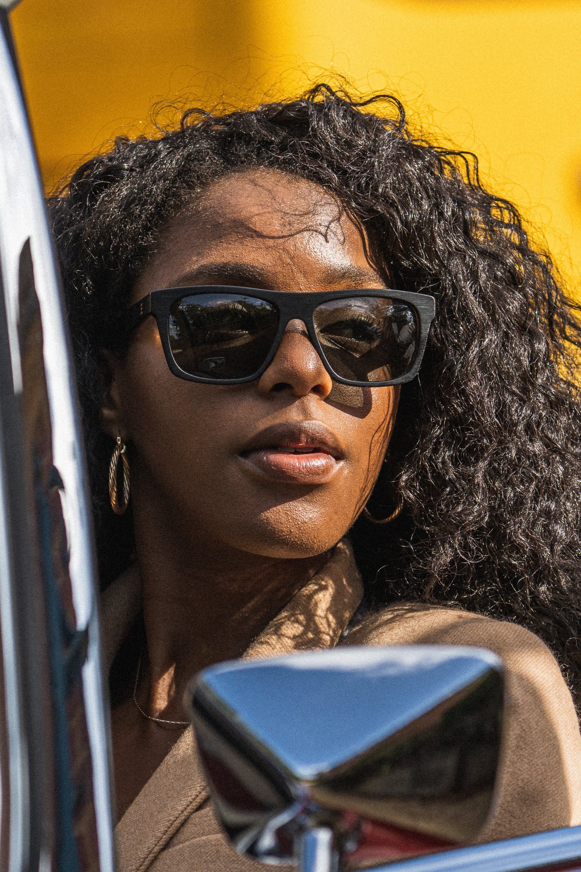 Woman with sunglasses, curly hair, in front of a yellow backdrop and car.