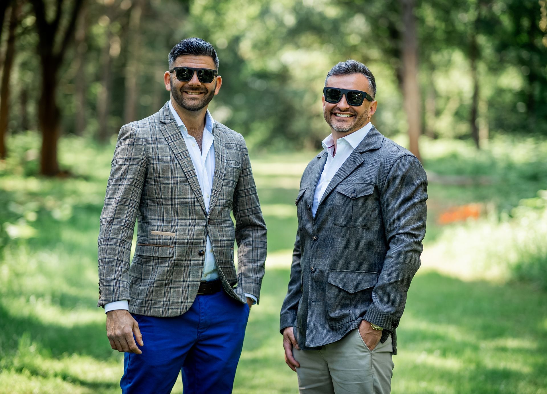 Two men in blazers and sunglasses standing outside in a park, smiling.