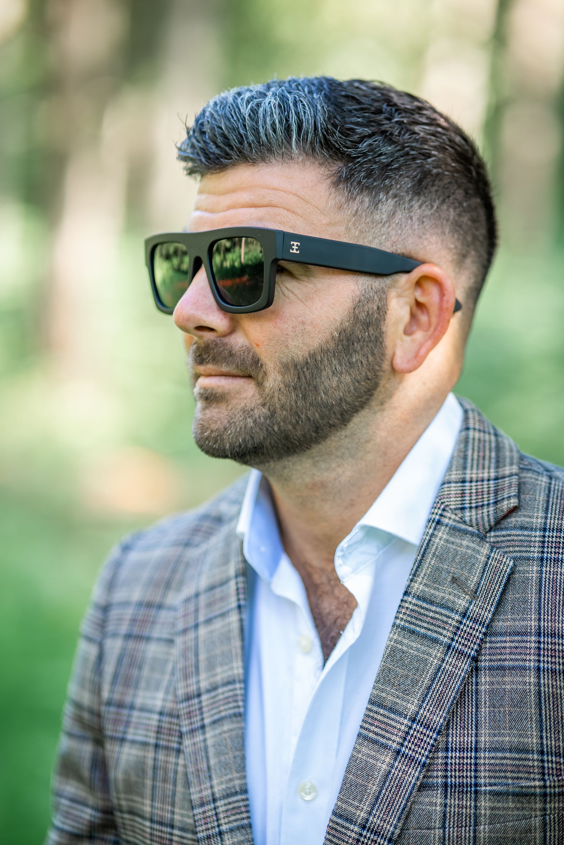 Man with beard wearing sunglasses and checkered jacket, outdoors in a green setting.