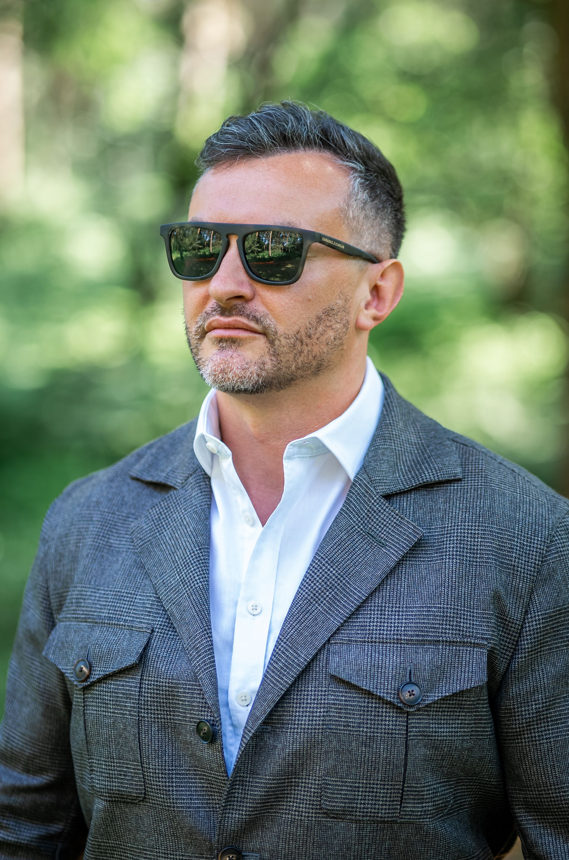 Man wearing sunglasses, gray jacket, white shirt, looking to the side, outdoors with green foliage.