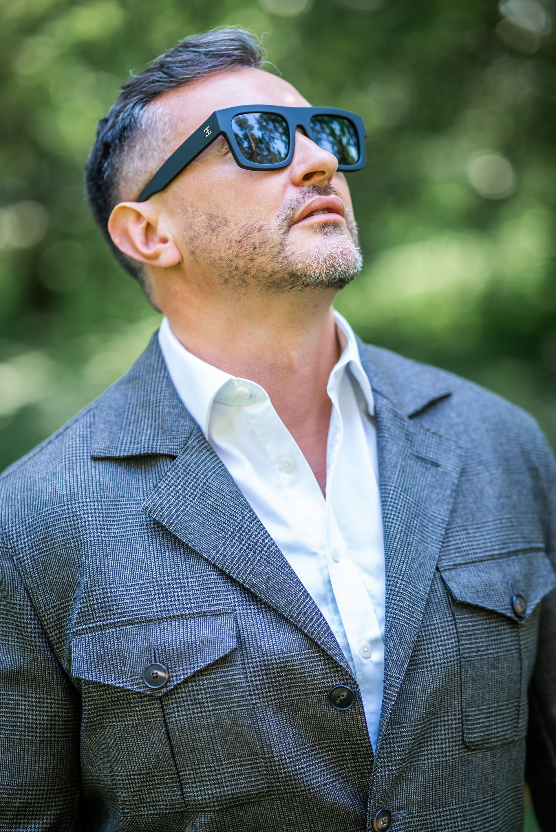 Man in sunglasses, looking upwards, wearing a textured gray jacket and white shirt outdoors.