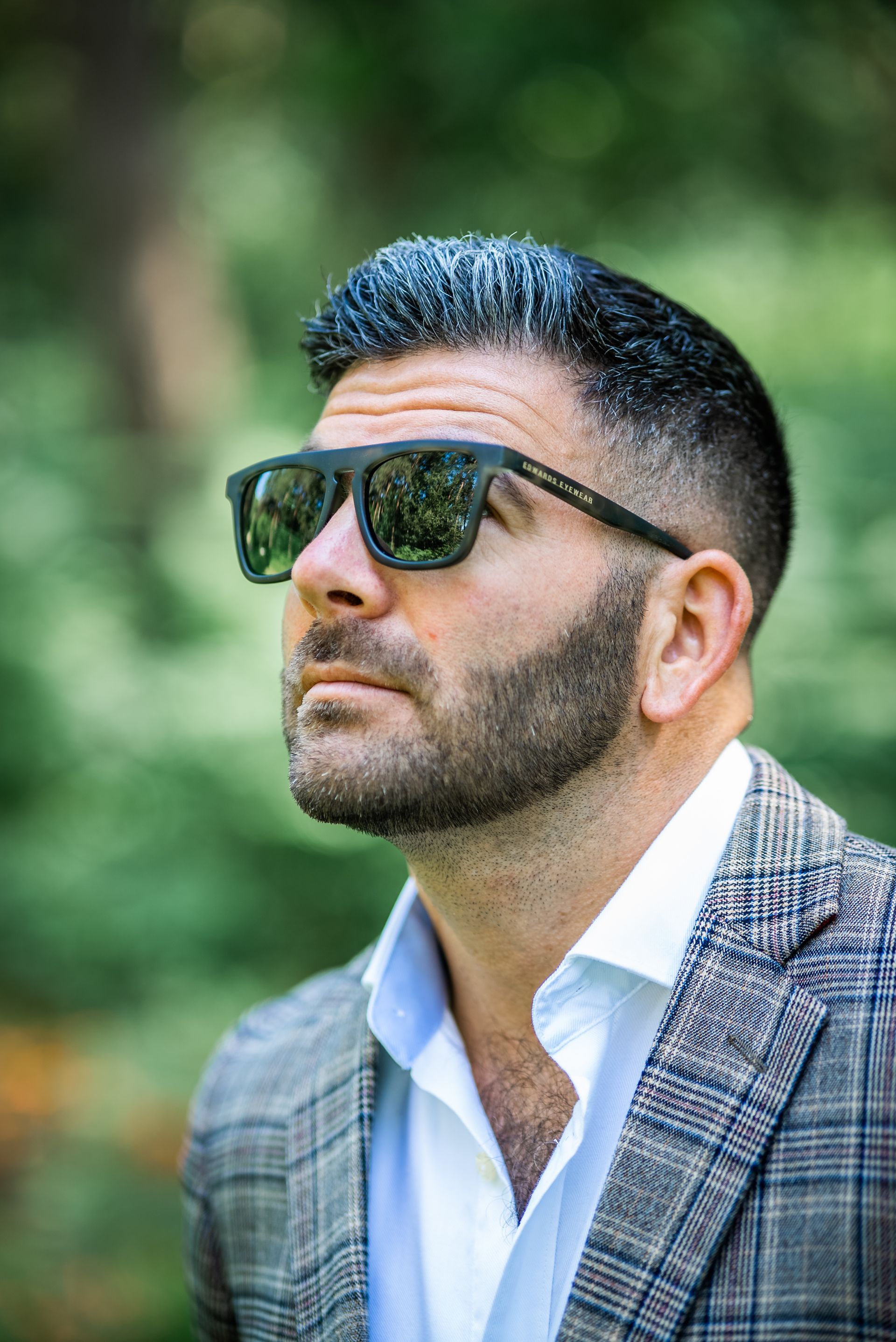 Man in sunglasses looking up; wearing a patterned blazer and white shirt, outdoors.