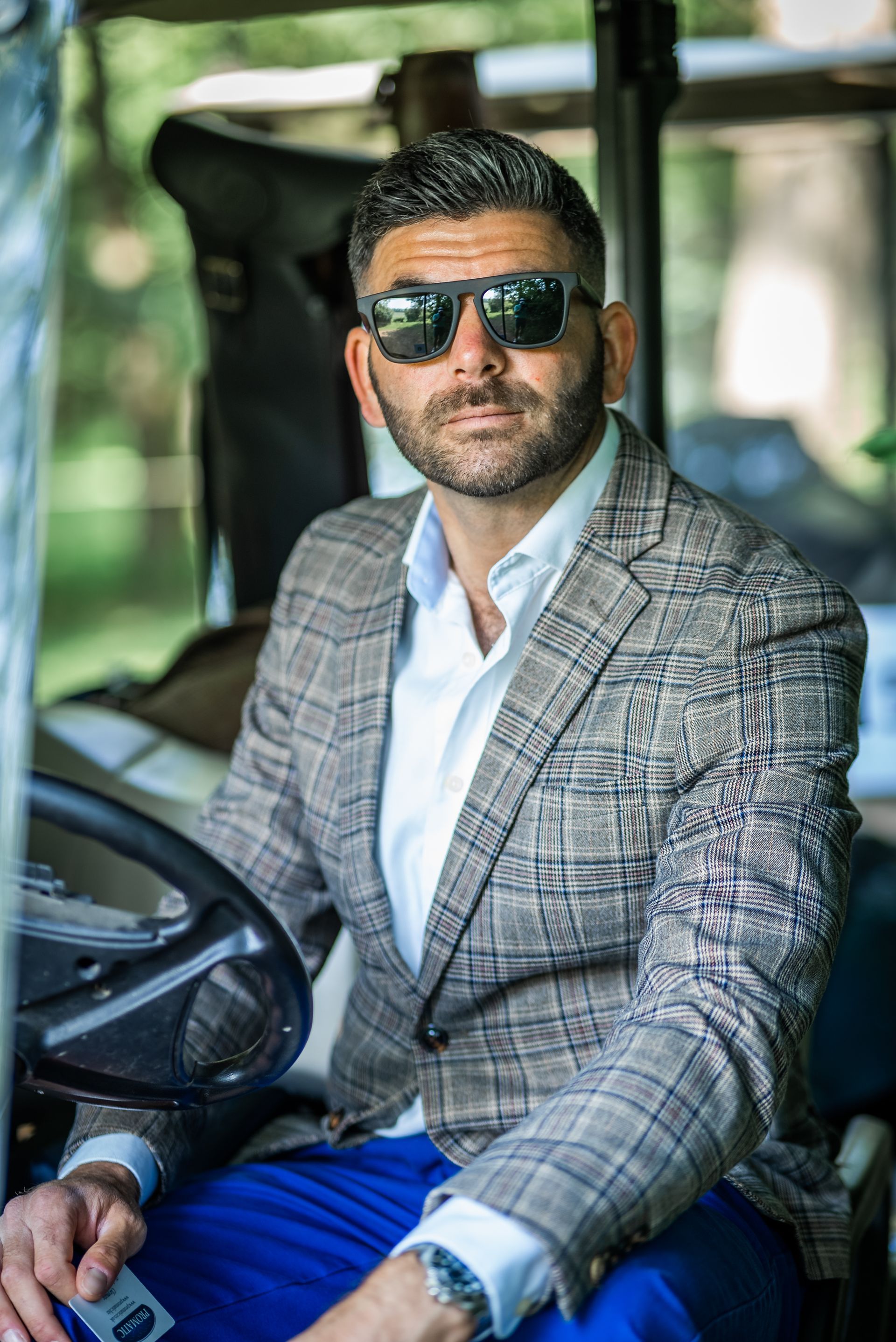 Man in sunglasses and plaid blazer sits in golf cart, smiling.