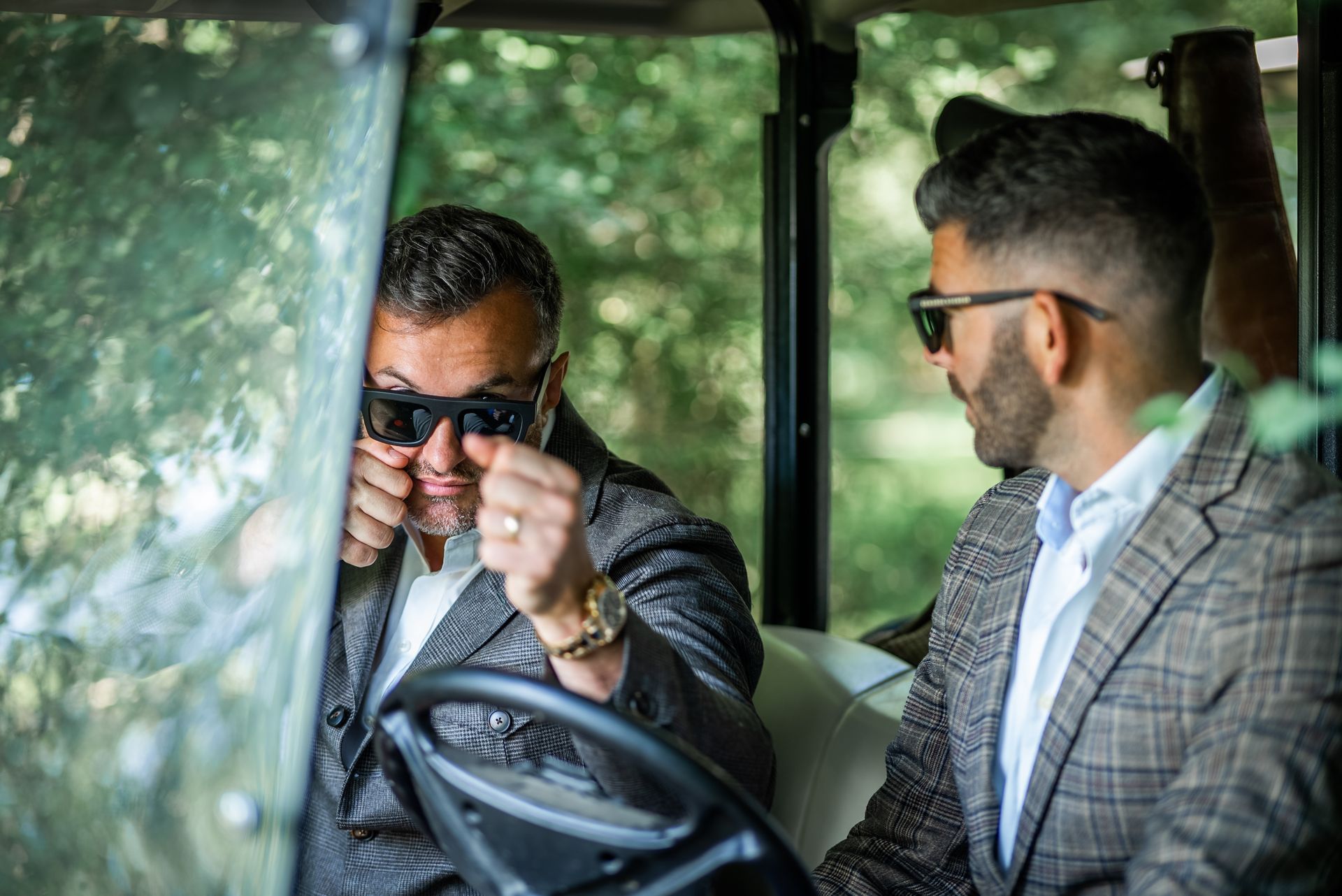 Two men in suits, wearing sunglasses, in a golf cart. One adjusts sunglasses while driving; the other looks on.