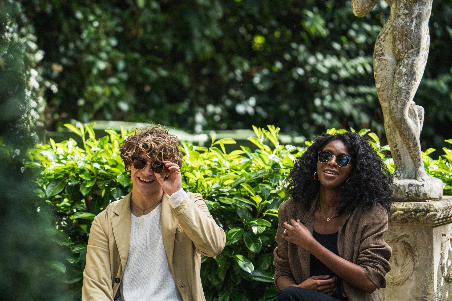 Two people in a garden, one looking through hands, the other wearing sunglasses, near a statue.