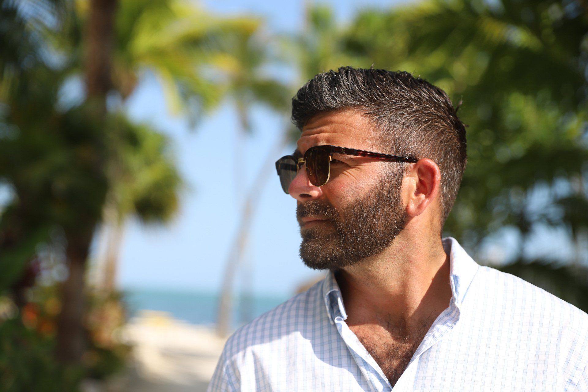 Man with beard and sunglasses outdoors, palm trees in background.