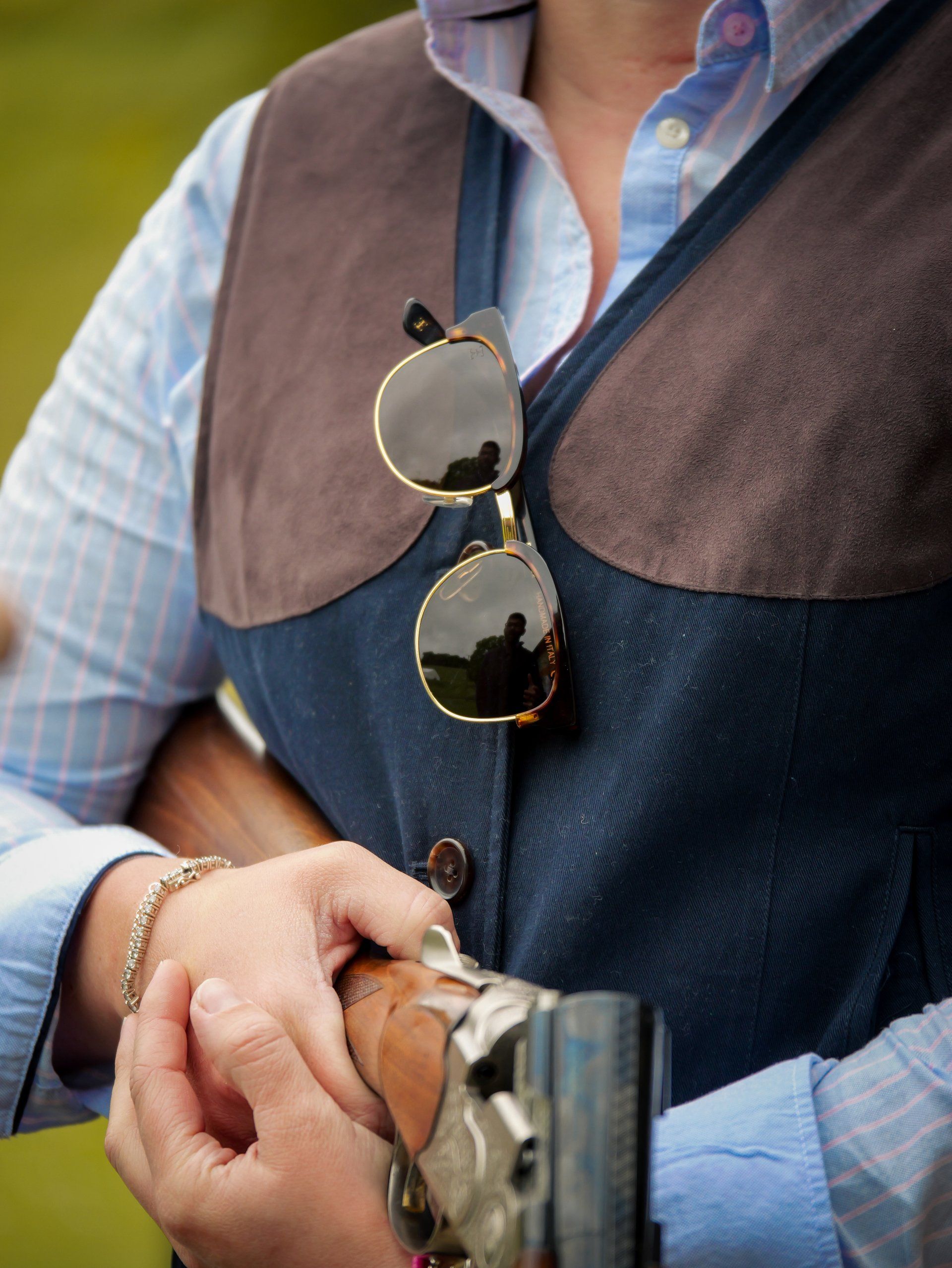 Woman holding a shotgun, wearing a vest with sunglasses clipped on. Outdoors.