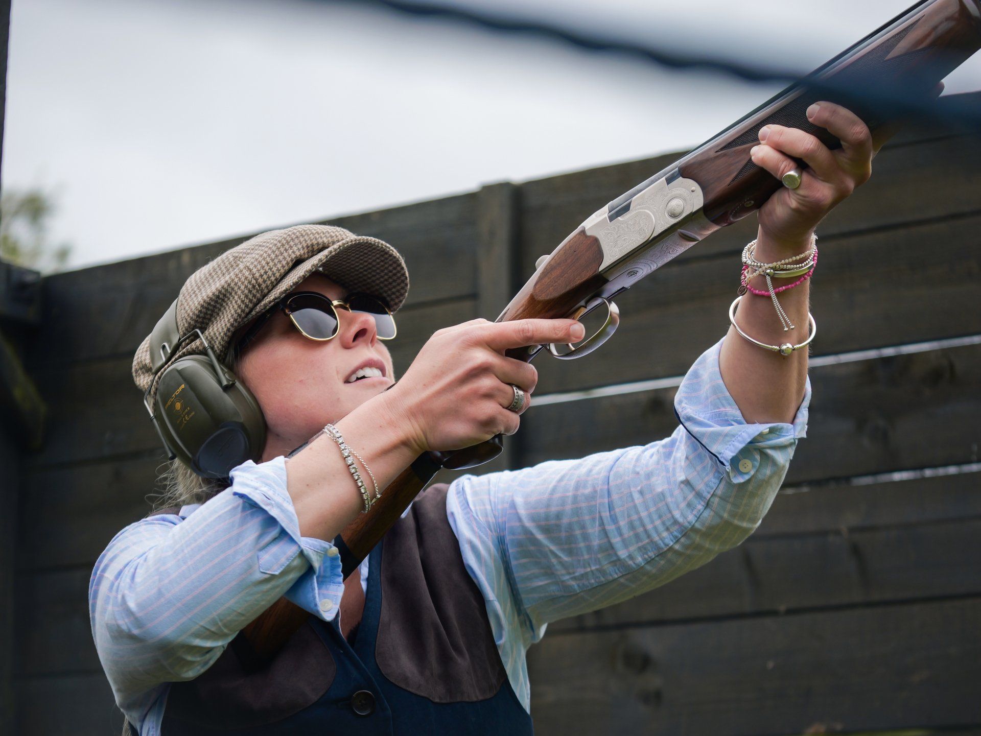 Woman shooting a shotgun, wearing ear protection, cap, sunglasses. Outdoors, aiming at the sky.