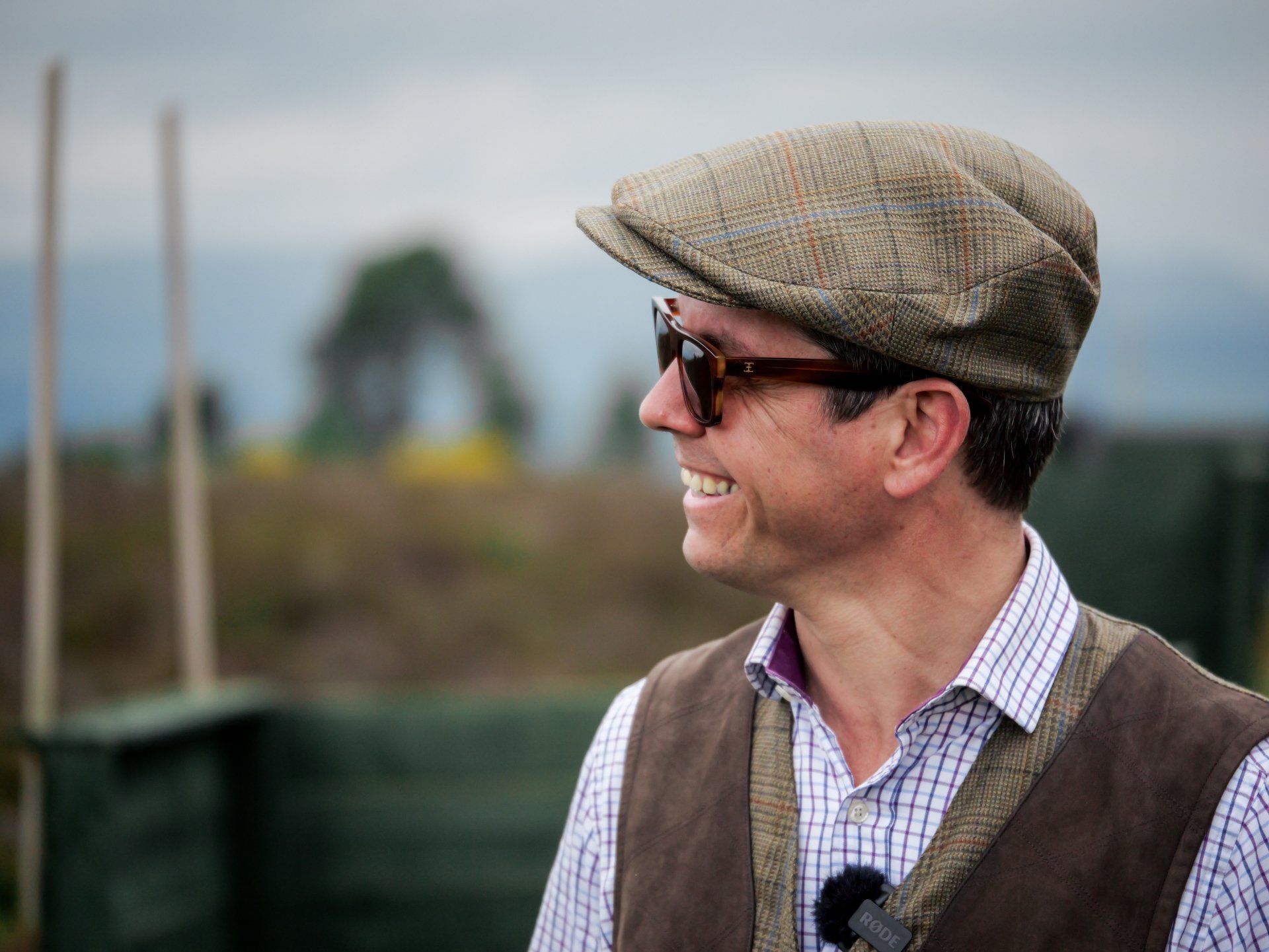 Man in tweed hat and sunglasses smiles outdoors, wearing a vest and collared shirt.